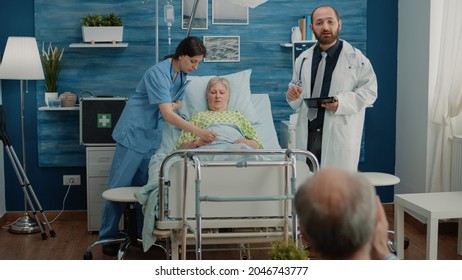 Medical Team Doing Examination For Sick Patient In Hospital Bed At Nursing Home Facility. Doctor Using Digital Tablet While Nurse Putting Oximeter On Hand Of Retired Woman For Checkup.