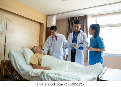 Medical Team On Rounds Meeting Around Bed Of Senior Male Patient At Hospital,Doctors Discussing The Report On Clipboard.