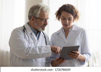 Medical Team Of Two Professional Doctors Talking And Using Digital Tablet. Senior Male Chief Physician Helping Young Female Nurse Holding Medical Tech Device Discussing Patient Diagnosis In Hospital.