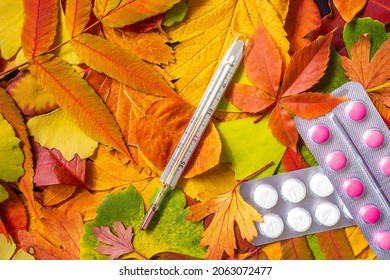 Medical Thermometer And Pills On A Background Of Fallen Leaves. High Temperature On Thermometer. Concept Of Autumn Common Cold, Covid Or Flu.