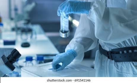 Medical Virology Research Scientist Works In A Hazmat Suit With Mask, She Inspects Test Tube With Isolated Virus String From Refrigerator Box. She Works In A Sterile High Tech Research Facility.