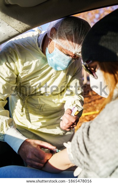 Medical worker doctor with protective equipment\
bouffant cap and face mask taking test blood sample with syringe\
and needle from patient in car in sunny day during corona-virus\
covid-19 pandemic
