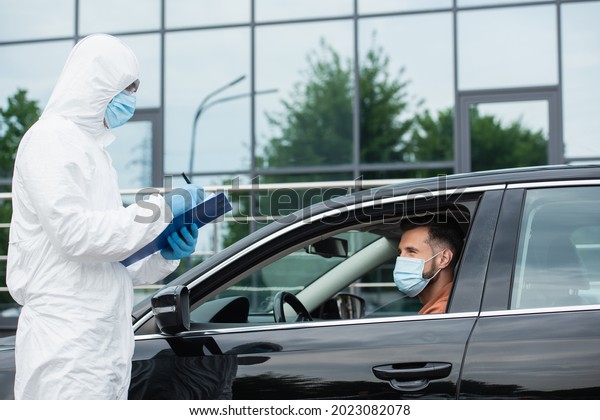 Medical worker in hazmat suit writing on clipboard\
near driver in medical\
mask