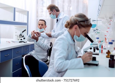 Medical Worker In Lab Coat And Sterile Mask, Doing A Microscope Analysis While Her Colleague Are Working Behind