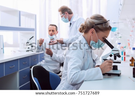 Medical worker in lab coat and sterile mask, doing a microscope analysis while her colleague are working behind