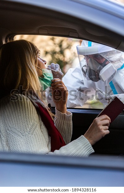 Medical worker performing drive-thru COVID-19\
test, taking nasal swab sample from female patient through car\
window, PCR diagnostic, doctor in PPE holding test kit. She is\
holding passports.
