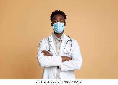 Medical Worker. Portrait Of Confident African American Doctor Wearing Protective Mask And Uniform Posing Over Beige Background In Studio, Black Male Physician Standing With Folded Arms, Copy Space