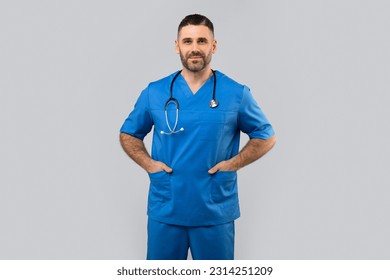 Medical worker. Portrait of confident middle aged doctor in blue uniform looking at camera while posing over light wall background. Cheerful therapist with stethoscope keeping hands in pockets - Powered by Shutterstock