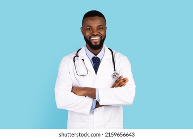 Medical Worker. Portrait Of Smiling African American Male Doctor In Uniform Standing With Folded Arms Over Blue Studio Background, Black Physician Wearing White Coat And Stethoscope, Copy Space