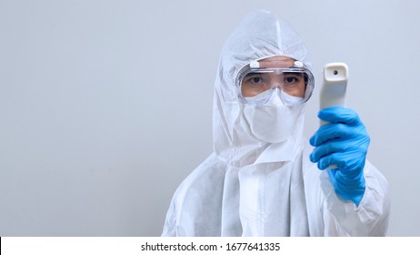 Medical Worker Scanning With Infrared Thermometer, IR Thermoscan, Confident Asian Doctor In Protective PPE Suit Wearing Face Mask And Eyeglasses