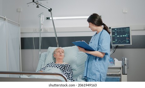 Medical worker taking notes about treatment progress while discussing with sick patient laying in hospital ward bed. Nurse giving assistance to retired woman with IV drip bag for healthcare - Powered by Shutterstock