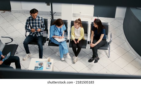 Medical Worker Talking To Little Girl And Mother In Waiting Area, Doing Healthcare Consultation With Patients At Hospital Reception Lobby. Nurse Examining Child In Facility Waiting Room.