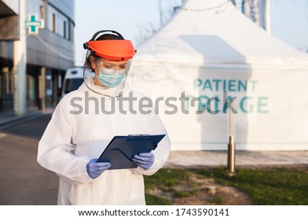 Medical worker wearing PPE protective white suit,face shield mask,holding clipboard patient health check up form,standing in front of clinic hospital,triage tent for COVID-19 UK point of care testing Foto stock © 
