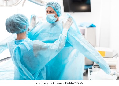 Medical Workers. Portrait Of Nurse In Sterile Gloves Putting On Blue Surgical Gown On Doctor