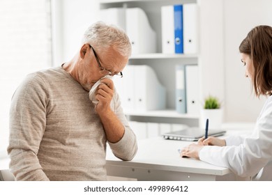 Medicine, Age, Health Care, Flu And People Concept - Senior Man Blowing Nose With Napkin And Doctor With Clipboard Writing At Medical Office At Hospital