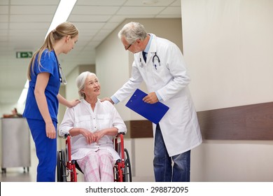 Medicine, Age, Health Care And People Concept - Doctor, Nurse And Senior Woman Patient In Wheelchair At Hospital Corridor