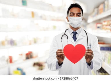 Medicine, Cardiology And Healthcare Concept - Indian Male Doctor Cardiologist Or Pharmacist Wearing Face Protective Medical Mask With Red Heart Shape And Stethoscope Over Drugstore Background