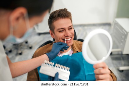 Medicine, dentistry and healthcare concept - closeup of a dentist with tooth color samples choosing a shade for a male patient's teeth in a dental clinic looking at a mirror.