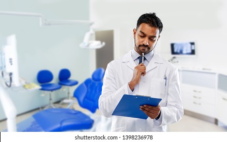 medicine, dentistry and profession concept - indian male dentist in white coat with clipboard over dental clinic office background
