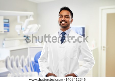 medicine, dentistry and profession concept - smiling indian male dentist in white coat over dental clinic office background
