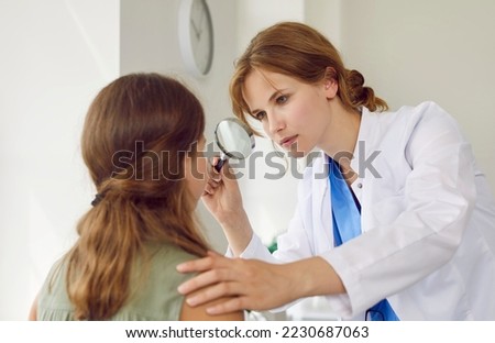 Medicine and dermatology. Professional dermatologist examines face of teenage girl with magnifying glass in her office. Young serious female doctor determines condition of skin of teenager's face.