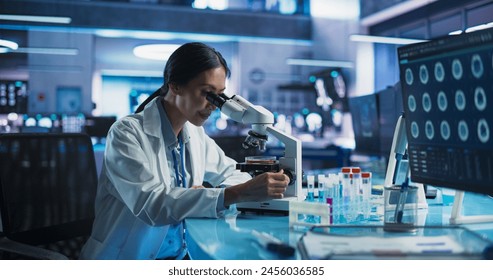 Medicine Development Laboratory: Asian Female Scientist Using Microscope, Analyzes Petri Dish Sample. Big Pharmaceutical Lab with Specialists Conducting Biotechnology Research, Developing New Drugs.