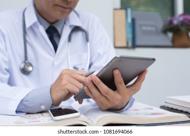 Medicine doctor with stethoscope holding pen, using digital tablet, reading anatomy textbook with smart phone on desk in doctor room. Medical student browsing internet tablet computer.