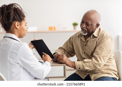 Medicine And Health Care Concept. Black Female Doctor In White Coat Holding Digital Tablet With Empty Screen For Mockup, Showing Male Patient His Test Results And Discussing Treatment, Sitting At Desk