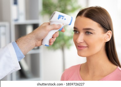 Medicine And Health Care Concept. Closeup Of Doctor With Contactless Infrared Themometer Doing Temperature Screening, Measuring Fever With Gun, Scanning Forehead Of Young Woman In Clinic