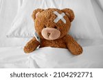 medicine, healthcare and childhood concept - ill teddy bear toy with medical patch on head and thermometer lying in bed