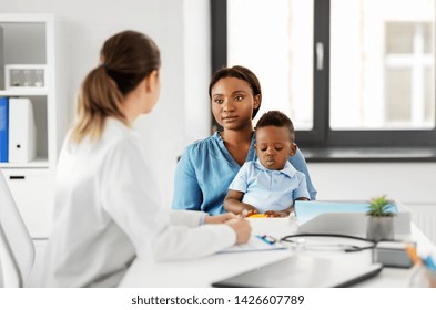 Medicine, Healthcare And Pediatry Concept - African American Mother With Baby Son And Caucasian Doctor At Clinic