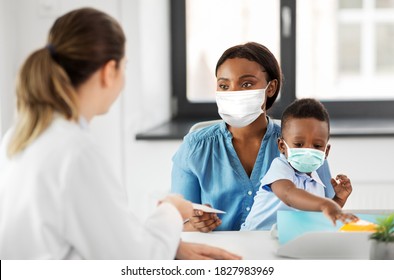 medicine, healthcare and pediatry concept - caucasian doctor giving prescription to african american mother wearing protective medical mask for protection from virus disease with baby son at clinic