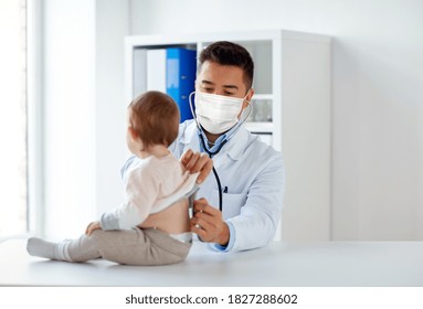medicine, healthcare, pediatry and people concept - doctor with stethoscope wearing face protective medical mask for protection from virus disease listening to baby on medical exam at clinic