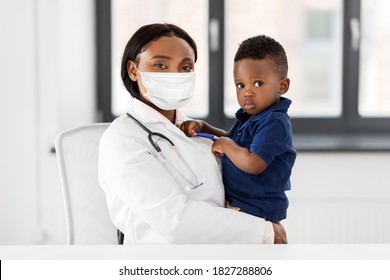 medicine, healthcare, pediatry and people concept - african american female doctor or pediatrician wearing protective mask holding baby boy patient on medical exam at clinic