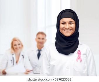 medicine, healthcare and people concept - smiling muslim female doctor wearing hijab and white coat with pink breast cancer awareness ribbon over clinic background - Powered by Shutterstock