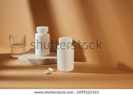 Medicine mockup for promotion, two white unbranded medicine bottle featured on brown background with a glass of water beside. Blank space for adding elements of designing, high angle shot