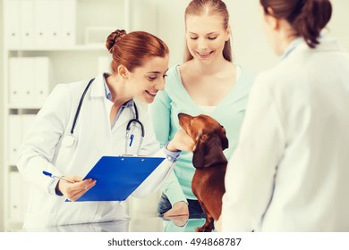 Medicine, Pet Care And People Concept - Happy Woman Holding Dachshund Dog And Veterinarian Doctor With Clipboard At Vet Clinic