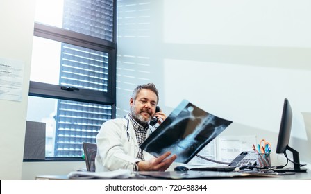 Medicine practitioner checking x-ray and using telephone in his office. Mature doctor examining x-ray and talking on phone.