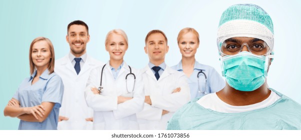 Medicine, Surgery And People Concept - Indian Male Doctor Or Surgeon In Mask, Goggles And Protective Wear Over Smiling Healthcare Workers Team On Blue Background