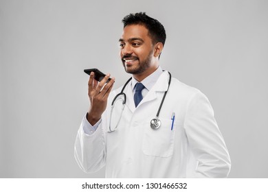 Medicine, Technology And Healthcare Concept - Smiling Indian Male Doctor In White Coat With Stethoscope Using Voice Command Recorder Over Grey Background