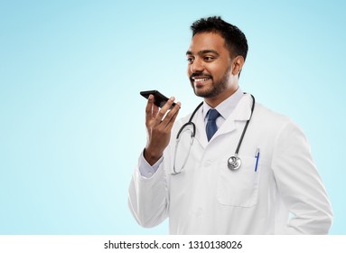 Medicine, Technology And Healthcare Concept - Smiling Indian Male Doctor In White Coat With Stethoscope Using Voice Command Recorder Over Blue Background