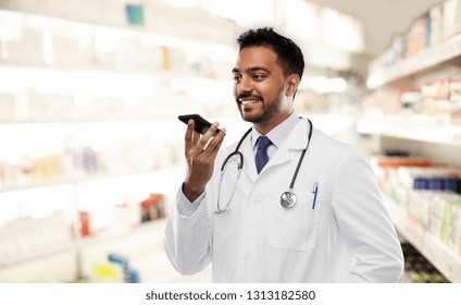 Medicine, Technology And Healthcare Concept - Smiling Indian Male Doctor Or Pharmacist In White Coat With Stethoscope Using Voice Command Recorder Over Drugstore Background