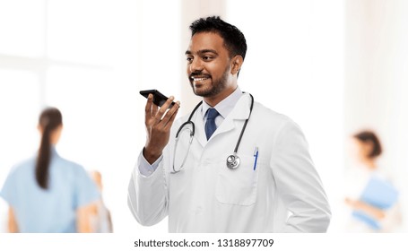 Medicine, Technology And Healthcare Concept - Smiling Indian Male Doctor In White Coat With Stethoscope Using Voice Command Recorder Over Hospital Background