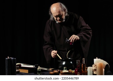medieval alchemist preparing magic potion while adding ingredient into pot isolated on black