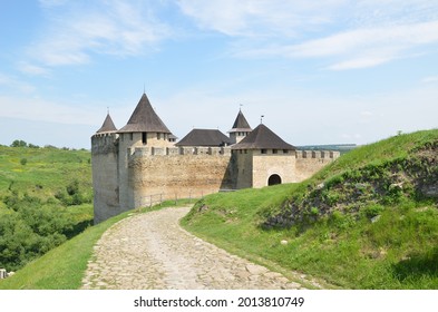 A medieval castle, with stone walls, loopholes, a wooden roof on the towers. The road leading to the castle is paved with cobble stones, surrounded by hills covered with green grass. - Shutterstock ID 2013810749