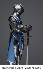 Medieval guard dressed in armor and blue surcoat holding a sword, standing in statue pose, against a gray background