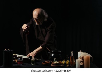 Medieval Healer Holding Flask And Reading Magic Cookbook Near Herbs Isolated On Black