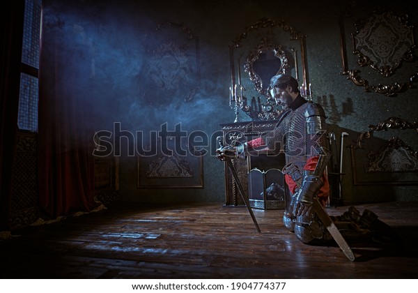 Medieval knight in armor knelt down and reads\
a prayer in the castle before the\
battle.