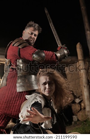 Medieval knight is going to make an execution of a witch. Witch house on the background.