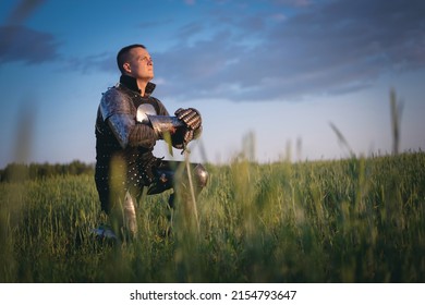 Medieval knight kneels in the field with his hand on the sword on a dark sky background. 